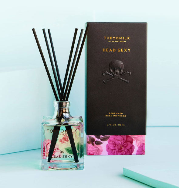 Black reeds emerge from a square glass bottle of TokyoMilk Dead Sexy diffuser oil next to its black and pink floral box packaging on a blue backdrop