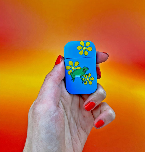 Model's hand wearing red nail polish holds a blue flip-top style lighter with yellow flowers and a green frog on it in front of a yellow-orange background