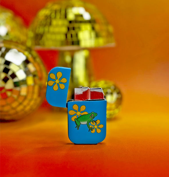 Blue lighter with yellow flowers and a green frog on it rests on a red-orange surface with its top flipped up to show the hardware inside; in the background are several gold disco ball mushroom props