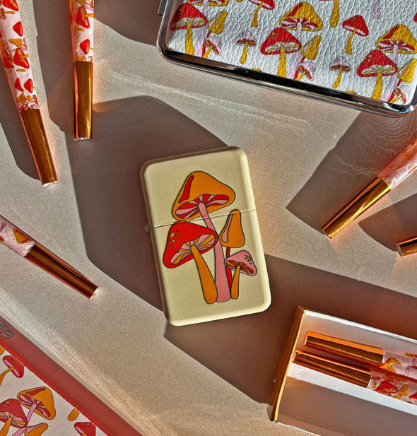 Cream-colored rectangular lighter with red, orange, and pink mushrooms design is staged with pre-rolled cigarettes and matching mushroom case