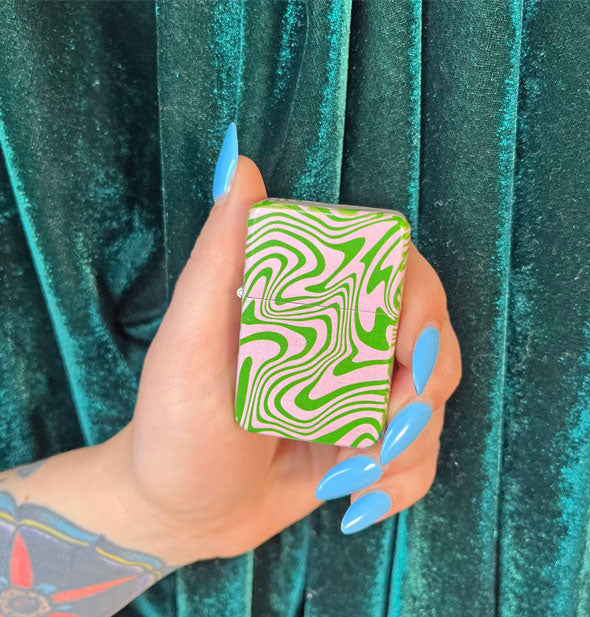 Model's hand with long blue fingernails holds a pink and green psychedelic swirl patterned reusable lighter against a green velvet backdrop