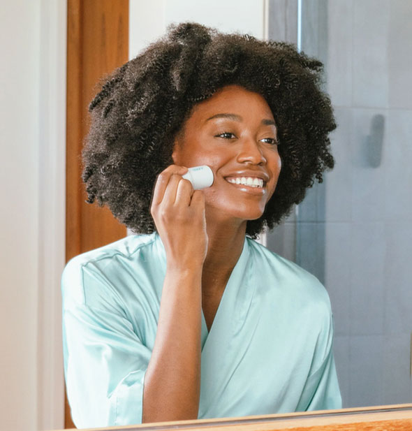 Mirror reflection of a smiling model applying Coola Refreshing Water Hydration Stick Sunscreen to cheek