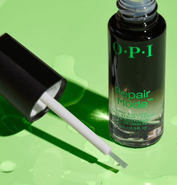Bottle and removed brush applicator lid of OPI Repair Mode Bond Building Nail Serum rest on a green surface dotted with moisture