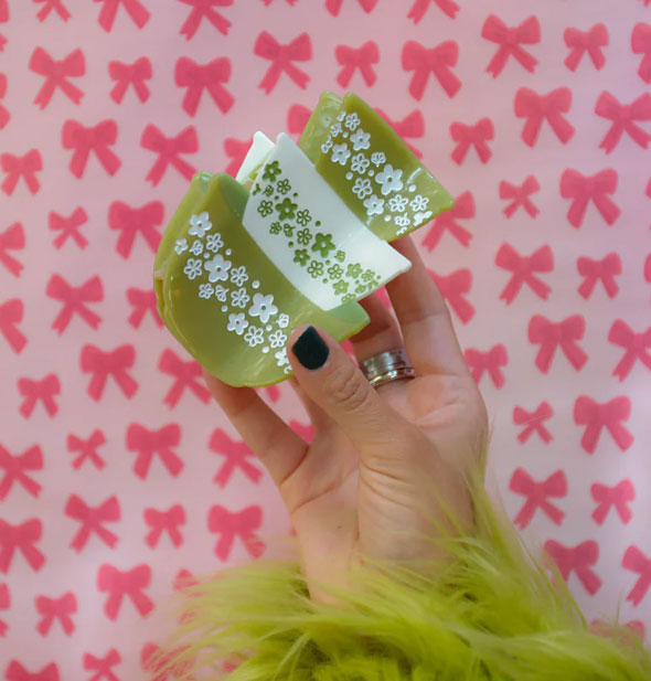 Model's hand holds a hair clip designed to resemble a stack of three green and white retro Pyrex bowls with daisy print against a pink bow patterned backdrop