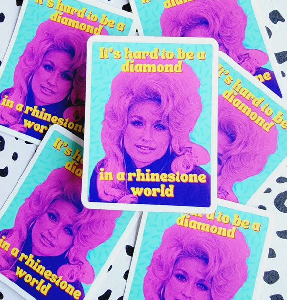 Pile of rectangular stickers on a black and white speckled surface feature a pink and purple image of Dolly Parton on a teal background and say, "It's hard to be a diamond in a rhinestone world" in yellow lettering