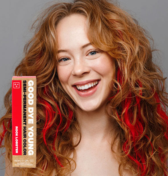 Model wears bright red streaks in wavy strawberry blonde hair; box of Good Dye Young Semi-Permanent Hair Color in shade Rock Lobster is inset at bottom left