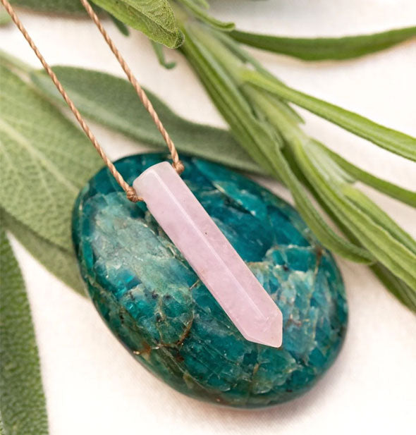 A pink rose quartz point necklace on gold cord rests on a smooth, round, green gemstone surrounded by green leaves