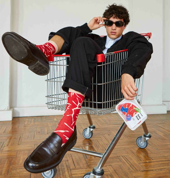 Model sits in a shopping cart with feet dangling over the edge wearing red and white Rump Steak crew socks with loafers