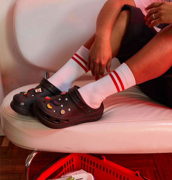 Model sitting on a white leather bench wears Crocs with white crew socks with double red stripe detail
