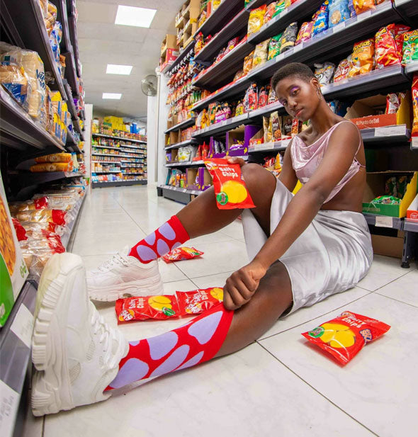 Model sitting on the floor in a convenience store aisle models a pair of Salty Chips Crew Socks and holds an unopened package in one hand