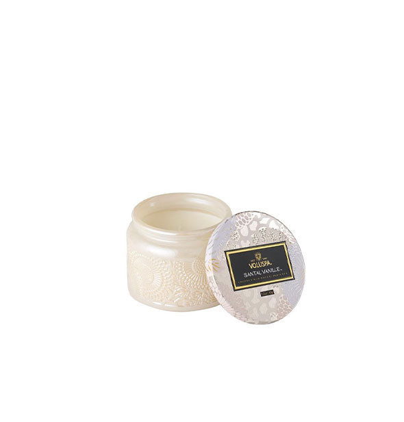 Frosted, opalescent white embossed glass Voluspa Santal Vanille candle jar with metallic white and silver floral tin lid propped alongside