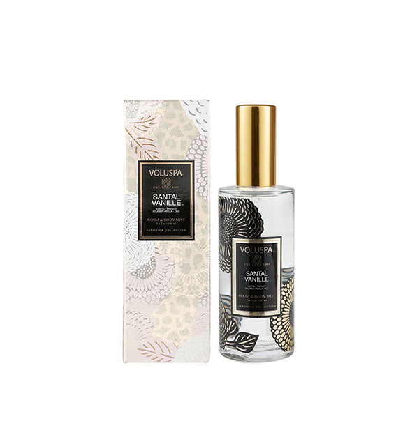 Floral-patterned clear glass bottle of Voluspa Santal Vanille Room & Body Mist next to metallic silvery gift box with white floral pattern