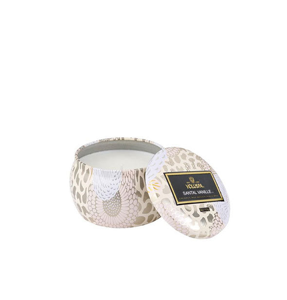 A small unlit candle inside a rounded tin with metallic floral design and matching lid set to the side