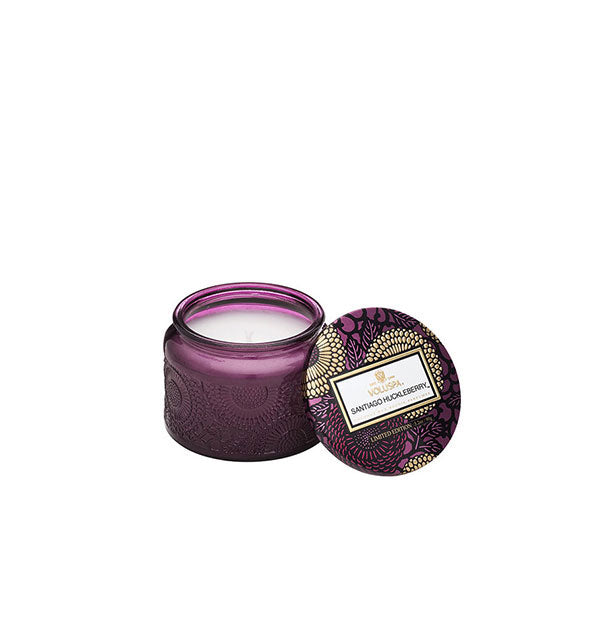 Embossed purple glass Santiago Huckleberry Voluspa candle jar with purple and gold metallic floral lid set to the side