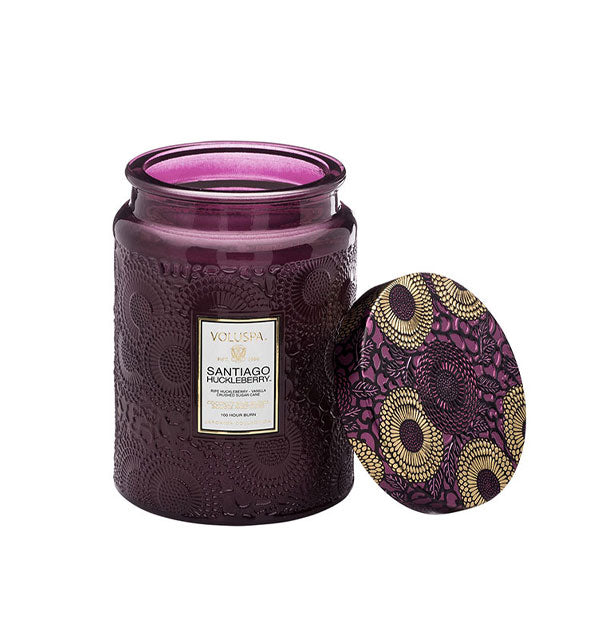 Purple embossed glass Santiago Huckleberry Voluspa candle jar with purple and gold metallic floral lid set to the side