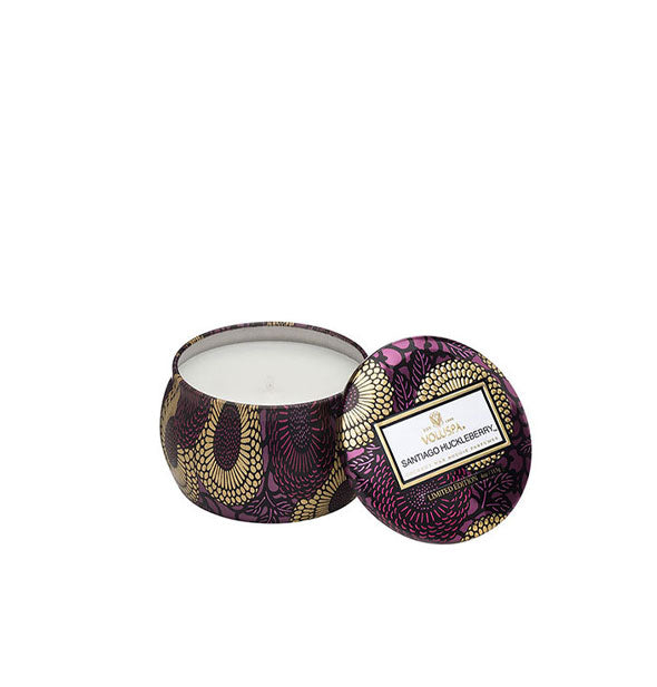 A small unlit candle inside a rounded tin with metallic purple and gold floral design and matching lid set to the side