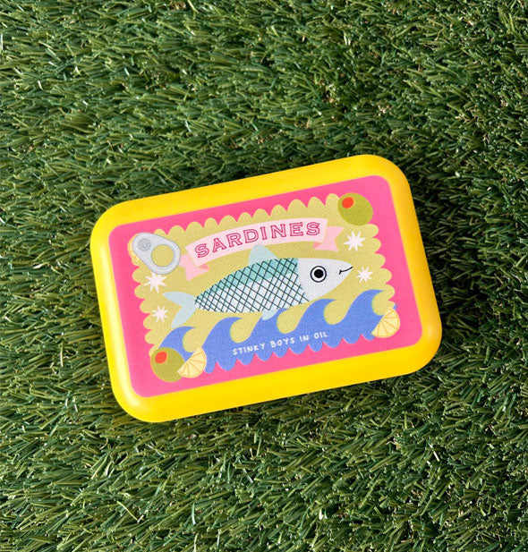 Rectangular yellow box with rounded corners and featuring a colorful illustration in the style of a can of sardines ("Stinky boys in oil") rests on green astroturf