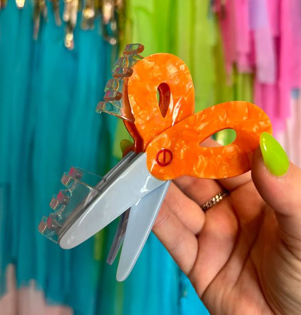 Model's hand holds a hair clip designed as a pair of orange-handled scissors in front of a colorful streamer backdrop