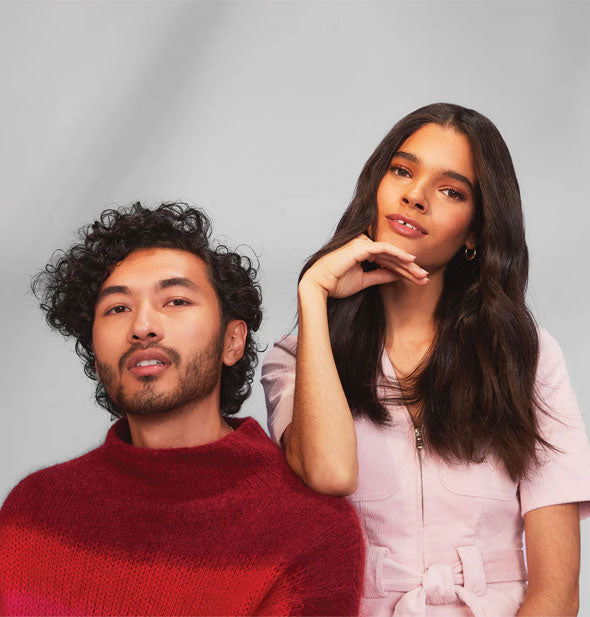 Model with long, sleek dark hair poses with elbow on the shoulder of another model with short, defined curls