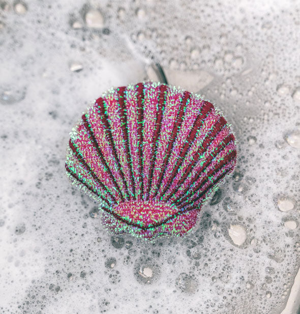 Closeup of a sparkly pink scallop seashell dish sponge resting in soap suds