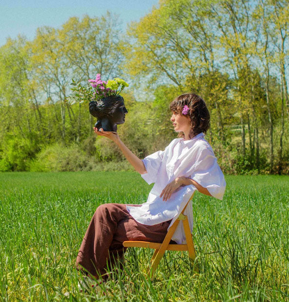Model sitting on a wooden folding chair in the middle of a tree-lined field holds a black Selene head vase in an outstretched right hand at eye level