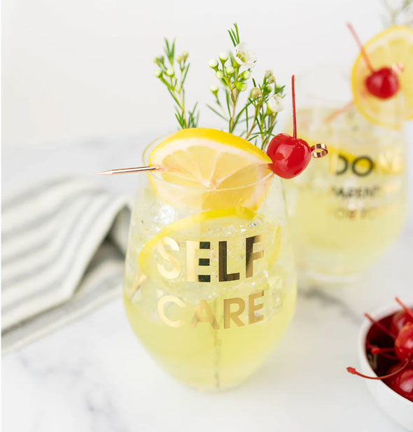 Self Care stemless wine glass is filled with a beverage and garnished with flowering herb sprigs, lemon slice, and maraschino cherry
