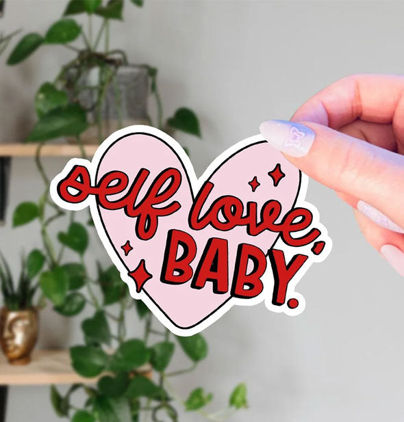 Model's hand holds a heart-shaped sticker overtop of which are the words, "Self love, Baby" in red lettering accented by red stars