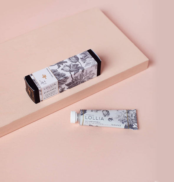 Mini tube and box of Lollia Elegance handcreme with black and white floral motif rest on a pink backdrop with small platform