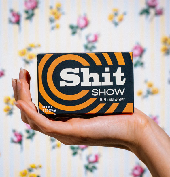 Model's hand holds a black and orange box of Shit Show Triple Milled Soap with radial stripe design and white lettering up in front of a floral backdrop