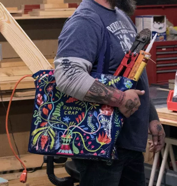 A tattooed model in a workshop holds the floral Random Crap Shopper filled with power tools over shoulder