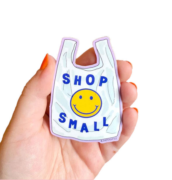 Model's hand holds a shopping bag-shaped sticker that says, "Shop Small" with smiley face graphic