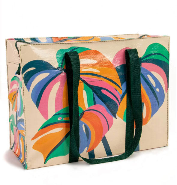 Cream-colored rectangular bag with dark green straps features illustrations of multicolored monstera leaves