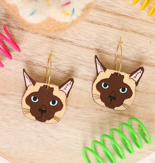 Pair of light and dark brown Siamese cat faces with blue eyes are on gold earring hoops resting on a wooden surface with brightly colored coils and a plush toy
