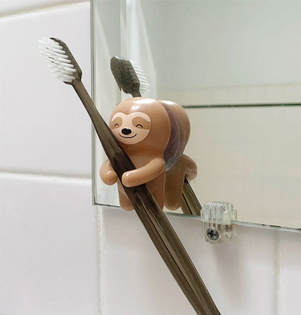 Smiling sloth toothbrush holder is suction-cupped to a bathroom mirror and holds a toothbrush between its arms