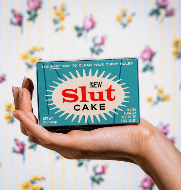 Model's hand holds a retro-looking box of New Slut Cake bar soap up in front of a floral backdrop