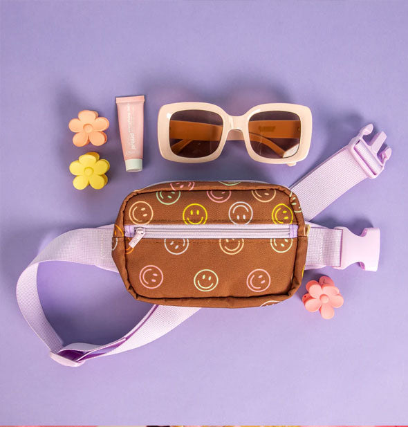 Brown and purple Small Smiley Hip Bag rests on a purple surface with flower clips, cosmetic tube, and a pair of square sunglasses