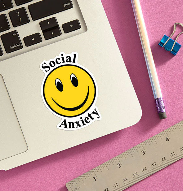 Social Anxiety smiley face sticker is applied to a laptop base panel and staged with pencil, ruler, and blue binder clip on a pink surface