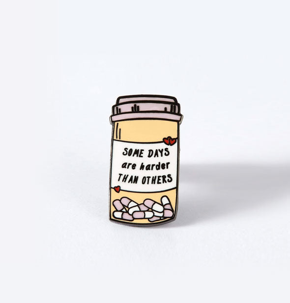 Enamel pin designed to resemble a prescription pill bottle says, "Some days are harder than others" on its label flanked by tiny red hearts