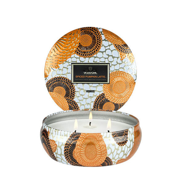 3-wick candle in a flat, rounded tin with black, orange, white, and metallic design accents and a matching lid standing up in the background