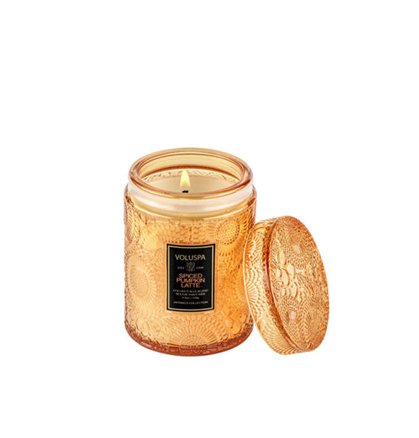 Burning candle in an orange embossed glass jar container with black label and matching lid set to the side