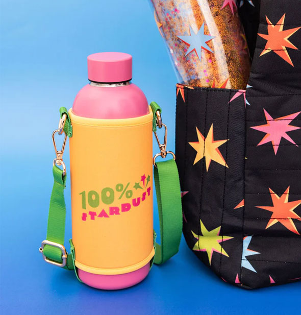 100% Stardust water bottle with sleeve and attached strap sits next to a black bag with colorful starburst pattern