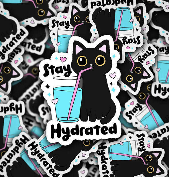 Pile of stickers featuring a wide-eyed black cat surrounded by small hearts and stars drinking water from a clear glass with pink straw say, "Stay Hydrated"