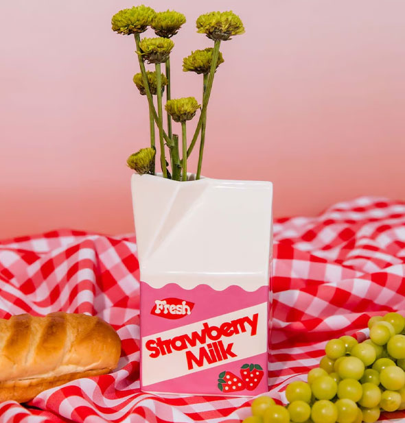 Strawberry milk carton vase holding a small bunch of flowers rests on a red gingham tablecloth with loaf of bread and green grapes