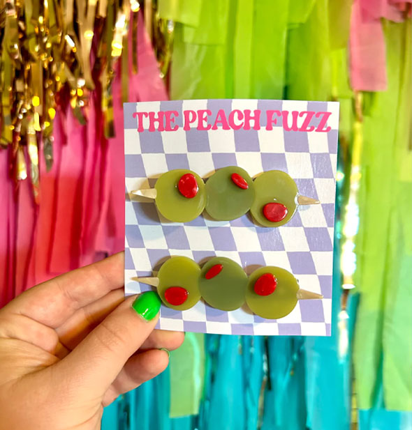 Model's hand holds a pair of olive trio hair clips on a purple and white checkered The Peach Fuzz product card in front of a colorful tinsel background
