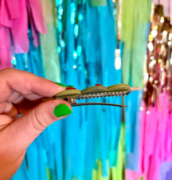 Model holds open the gold alligator-style clip hardware of an olive trio hair clip in front of a colorful tinsel backdrop
