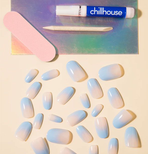 A smattering of pastel blue-to-pink ombre press-on nails with Chillhouse glue tube, pink file, and wooden cuticle pusher on an iridescent tray