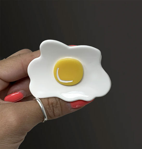 Model's hand holds a white and yellow sunny side up hair claw clip against a dark background