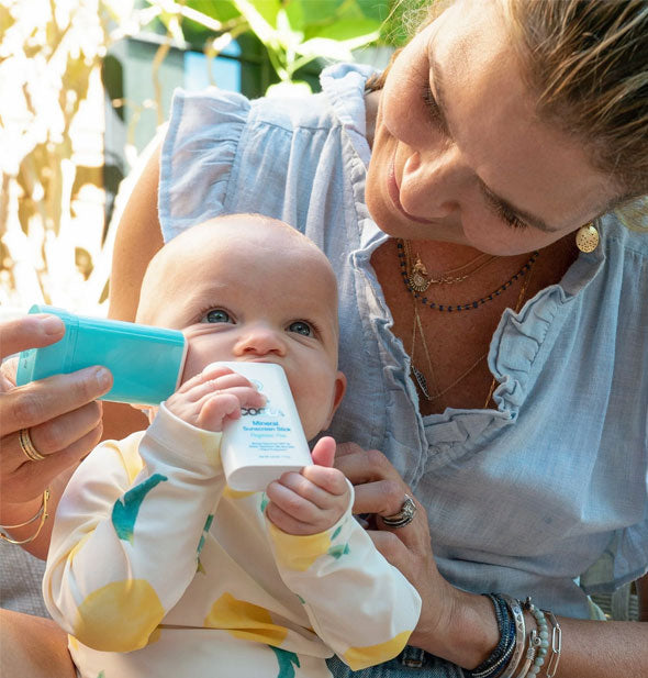 Model applies Coola Mineral Sunscreen Stick to baby's cheek while the baby nibbles on the lid
