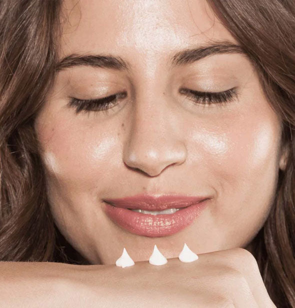 Model looks down at three dollops of hand cream applied to back of hand