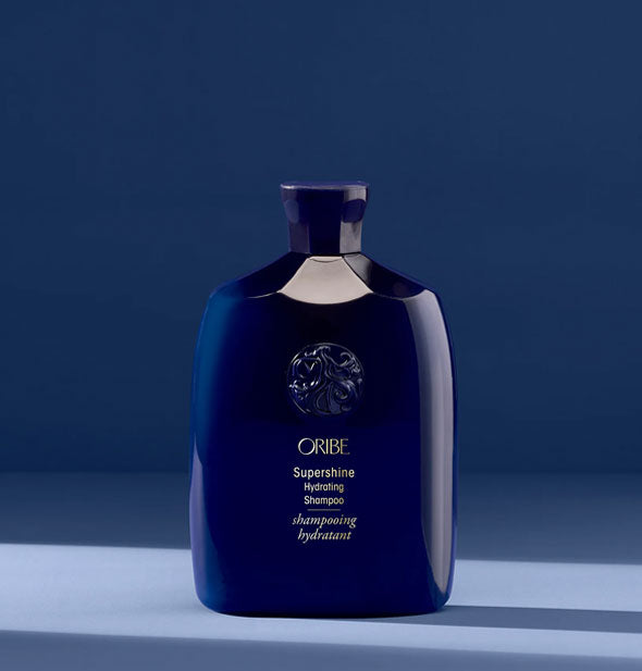 Dark blue 8.5 ounce bottle of Oribe Supershine Hydrating Shampoo on a dark blue shadowy backdrop